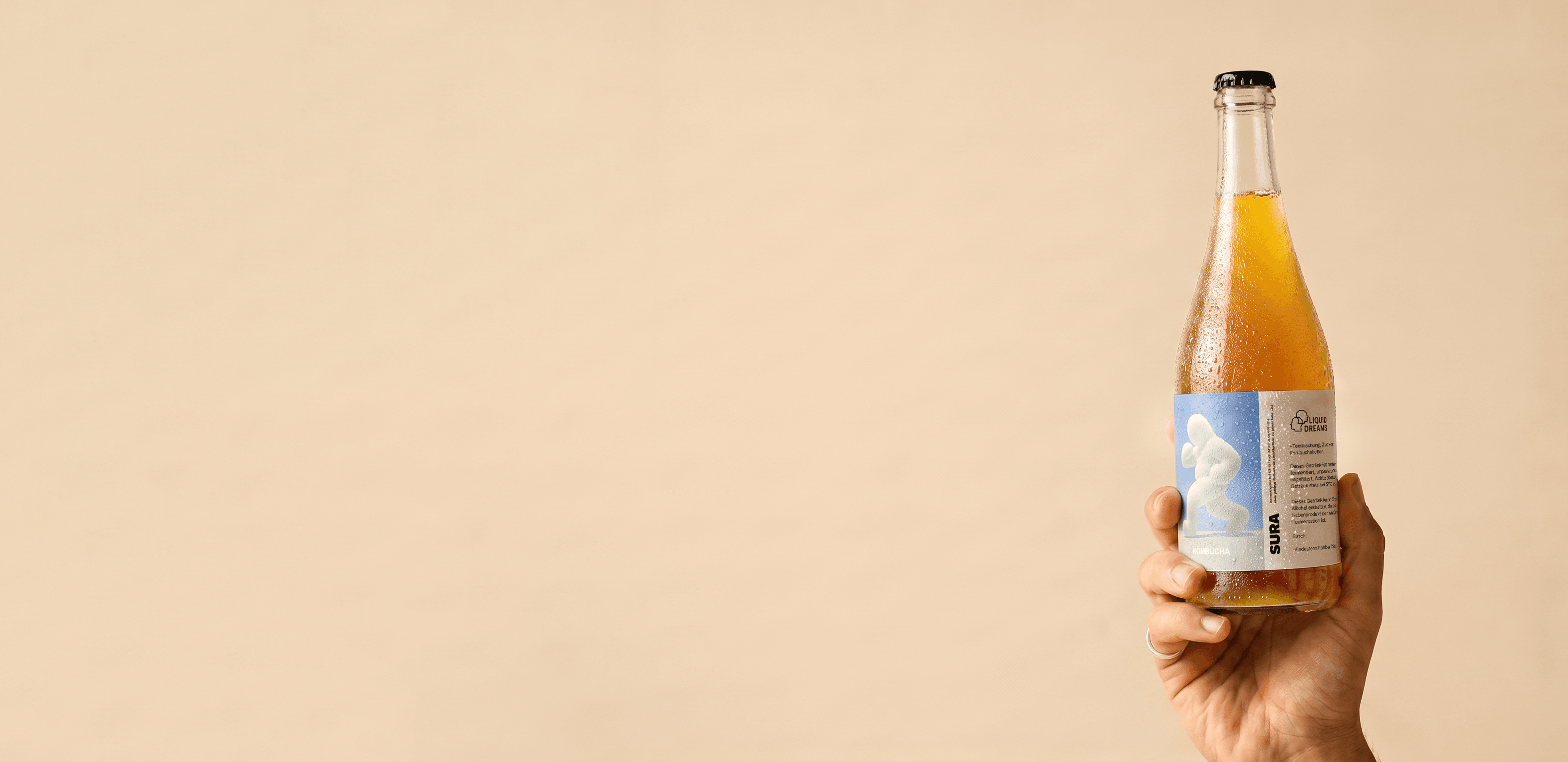 A hand holding a Liquid Dreams SURA kombucha bottle aloft against a warm beige studio backdrop, generous negative space to the left