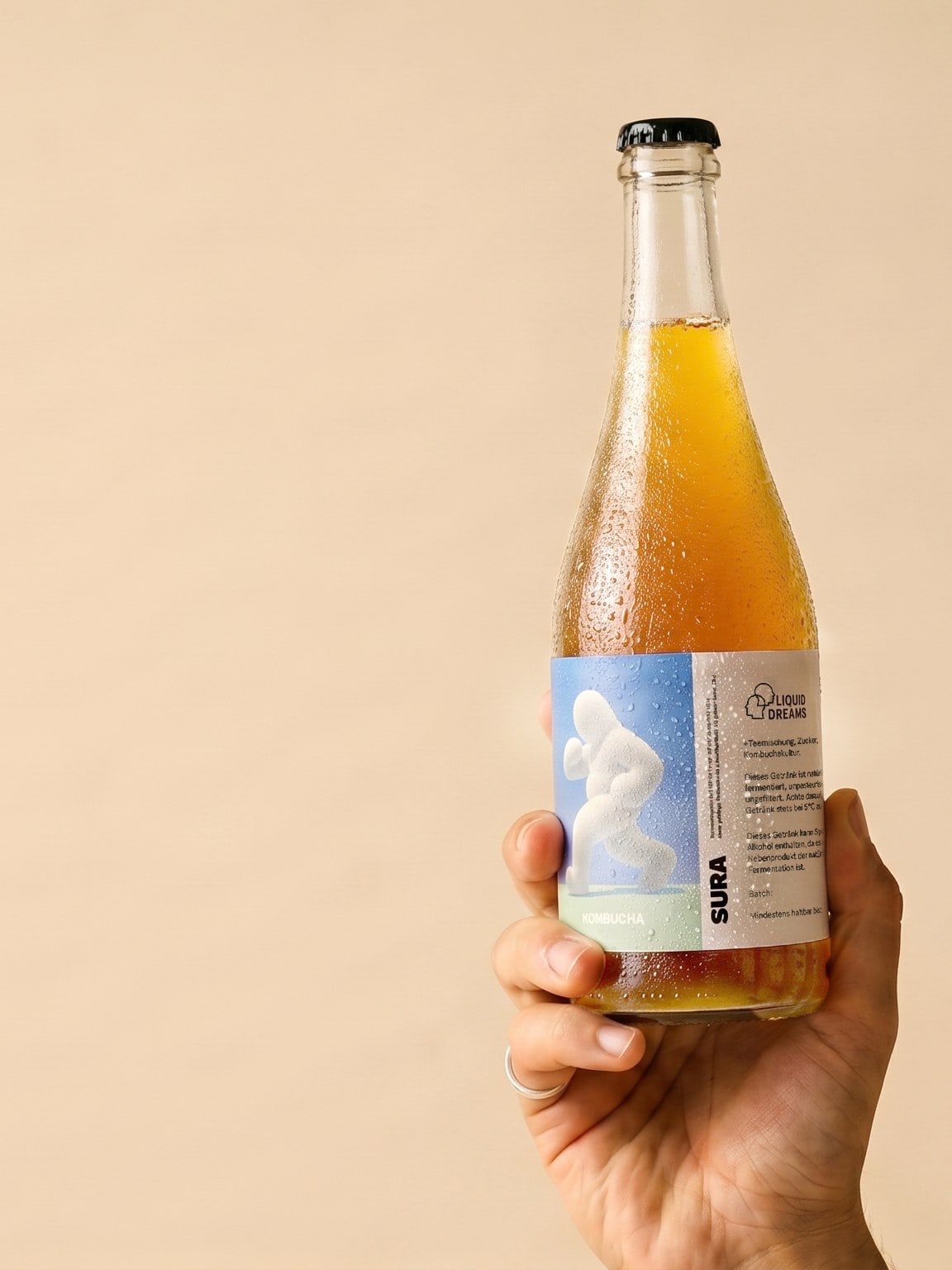 A hand holding a Liquid Dreams SURA kombucha bottle against a warm beige backdrop