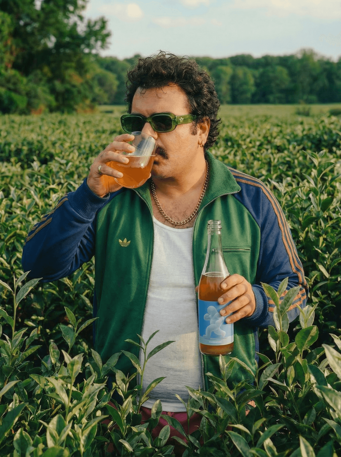 A man in a green tracksuit and sunglasses drinking SURA from a wine glass in a tea field, bottle in hand